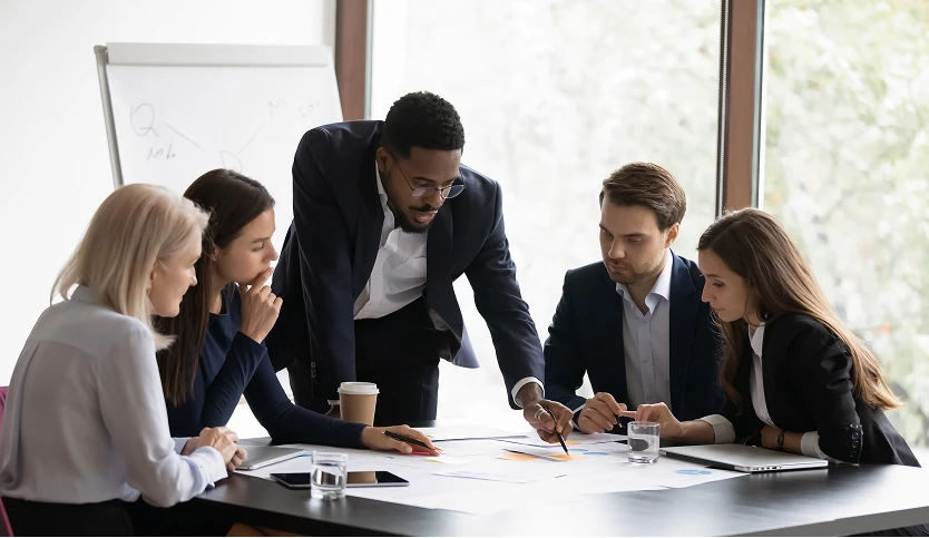 Man in a suit pointing to documents spread out on a table during a meeting, representing professional collaboration, strategy discussion, or document review in a business setting